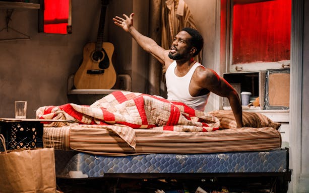 Man sitting on bed in a cluttered room with a guitar in the background, Topdog/Underdog theme.