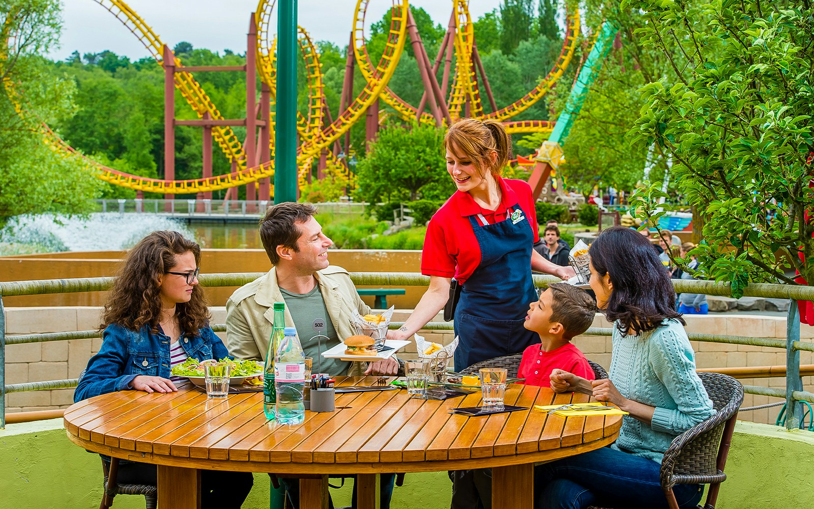 Family dining outdoors with roller coaster in background at Parc Asterix, France.