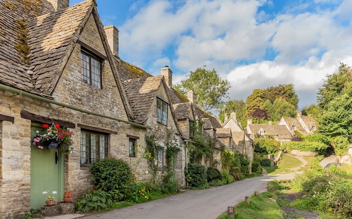 Cotswolds stone cottages along a village road, lush greenery, and blue sky.