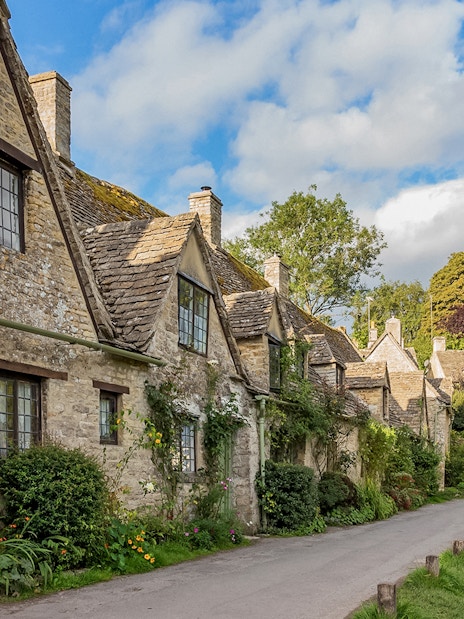 Cotswolds stone cottages along a village road, lush greenery, and blue sky.