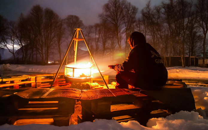 Person warming hands by campfire at Tromsø safari camp during aurora chase.