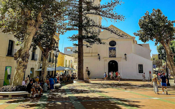 Church and plaza in Nerja's old town with people walking and sitting nearby.