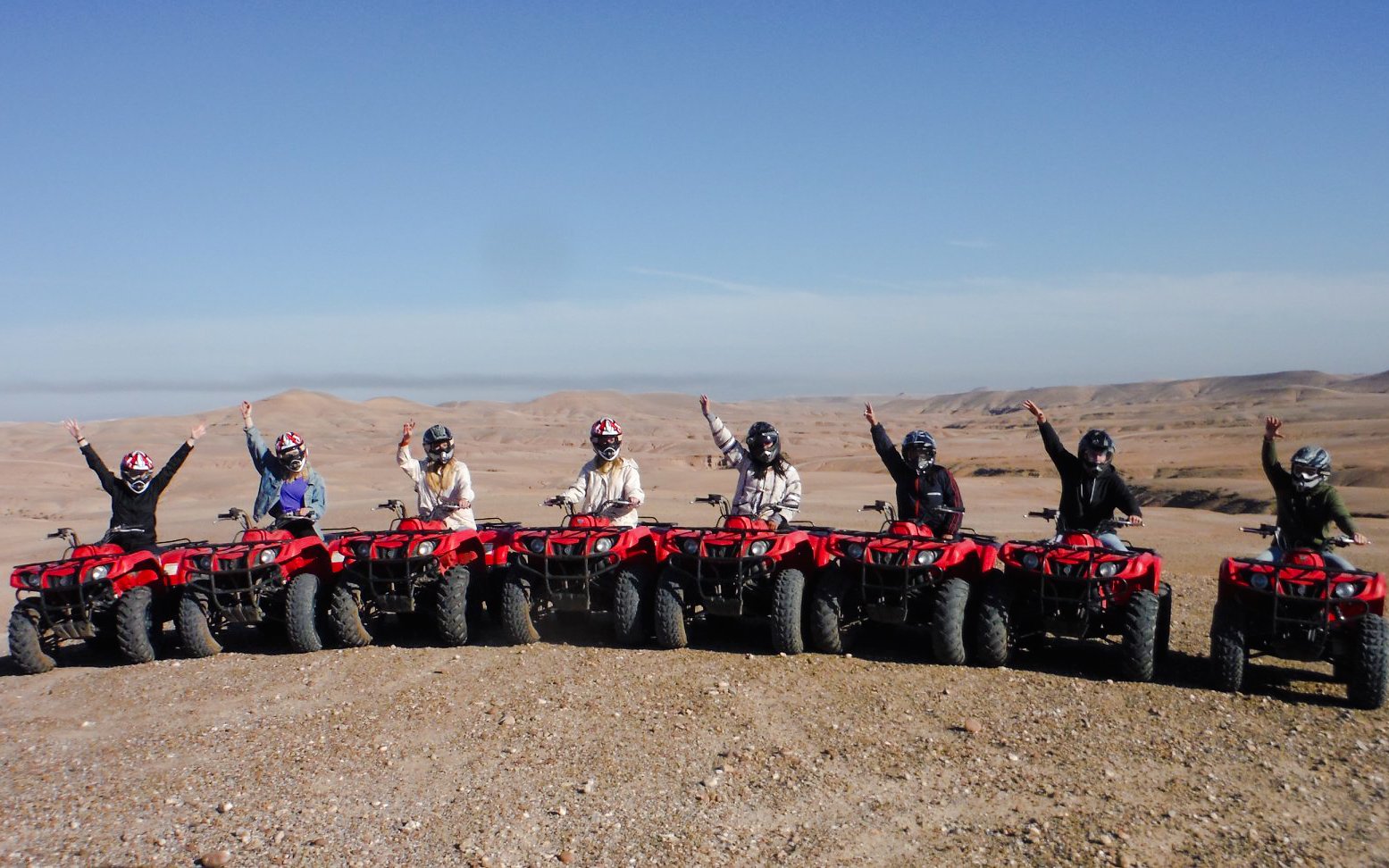 Group on red quad bikes in Agafay Desert, Morocco, raising arms in excitement.
