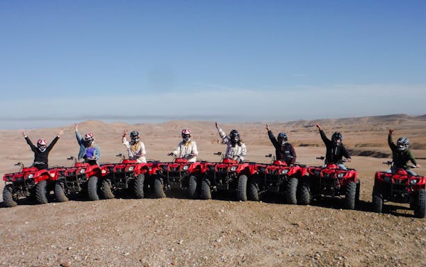Group on red quad bikes in Agafay Desert, Morocco, raising arms in excitement.