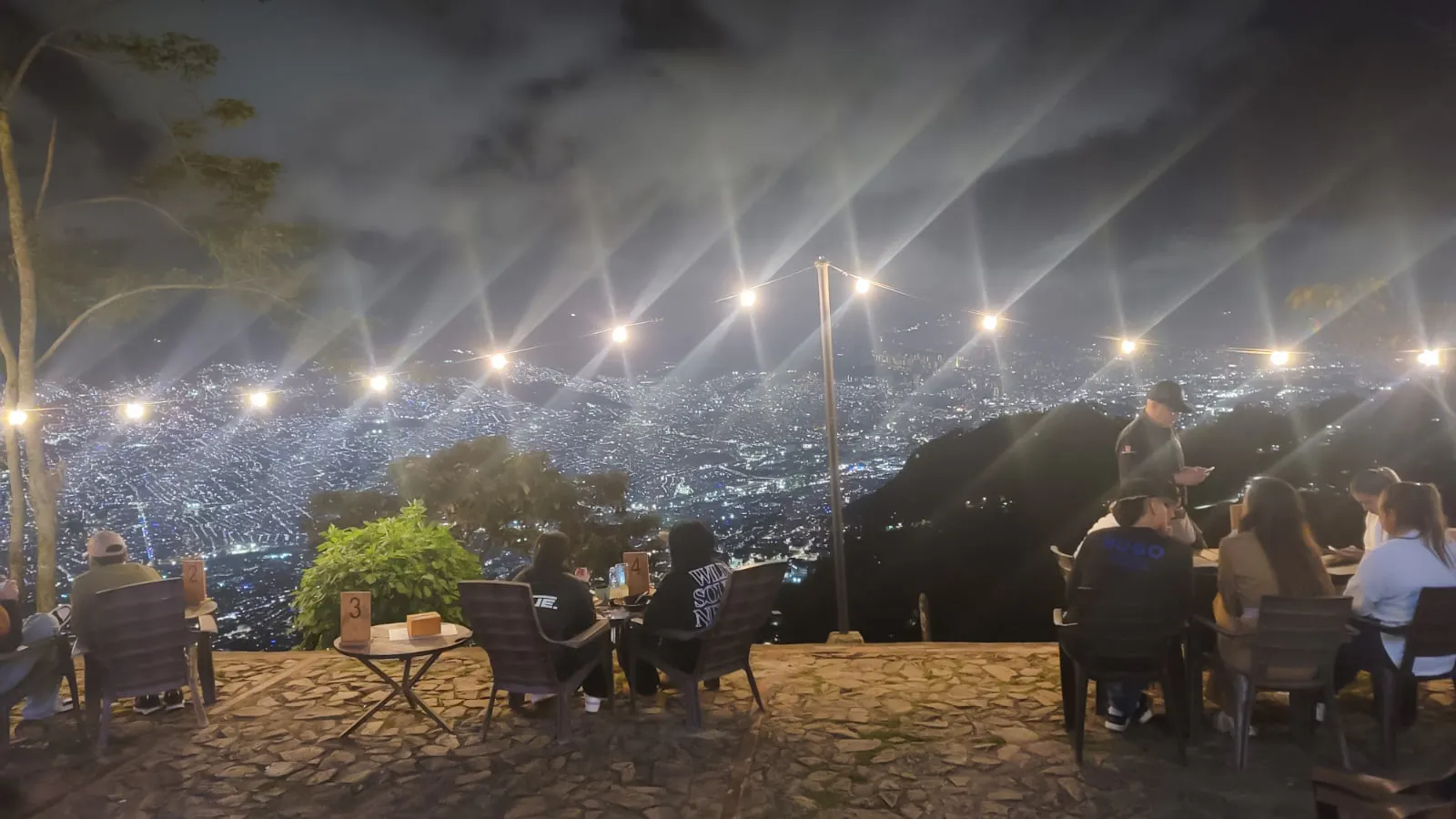 Night view of Medellin from a hillside restaurant with diners enjoying local food.