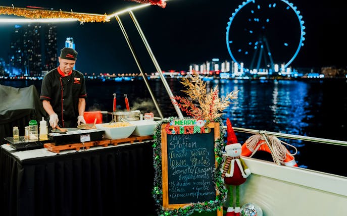 Chef preparing pasta on a Christmas dinner cruise with a view of a lit-up Ferris wheel at night.