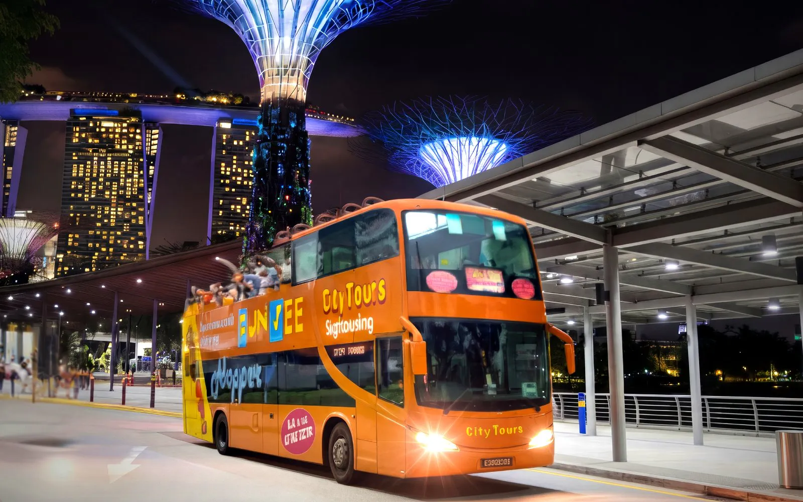 Open-top tour bus in Singapore at night with Marina Bay Sands and Supertree Grove in the background.