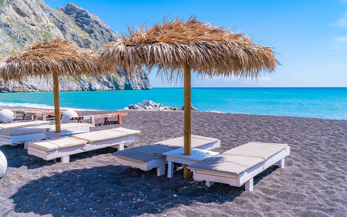 Sun loungers under straw umbrellas on a black sand beach in Santorini, Greece.