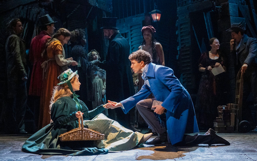 Les Misérables stage scene with actors in period costumes, one kneeling and offering a hand.
