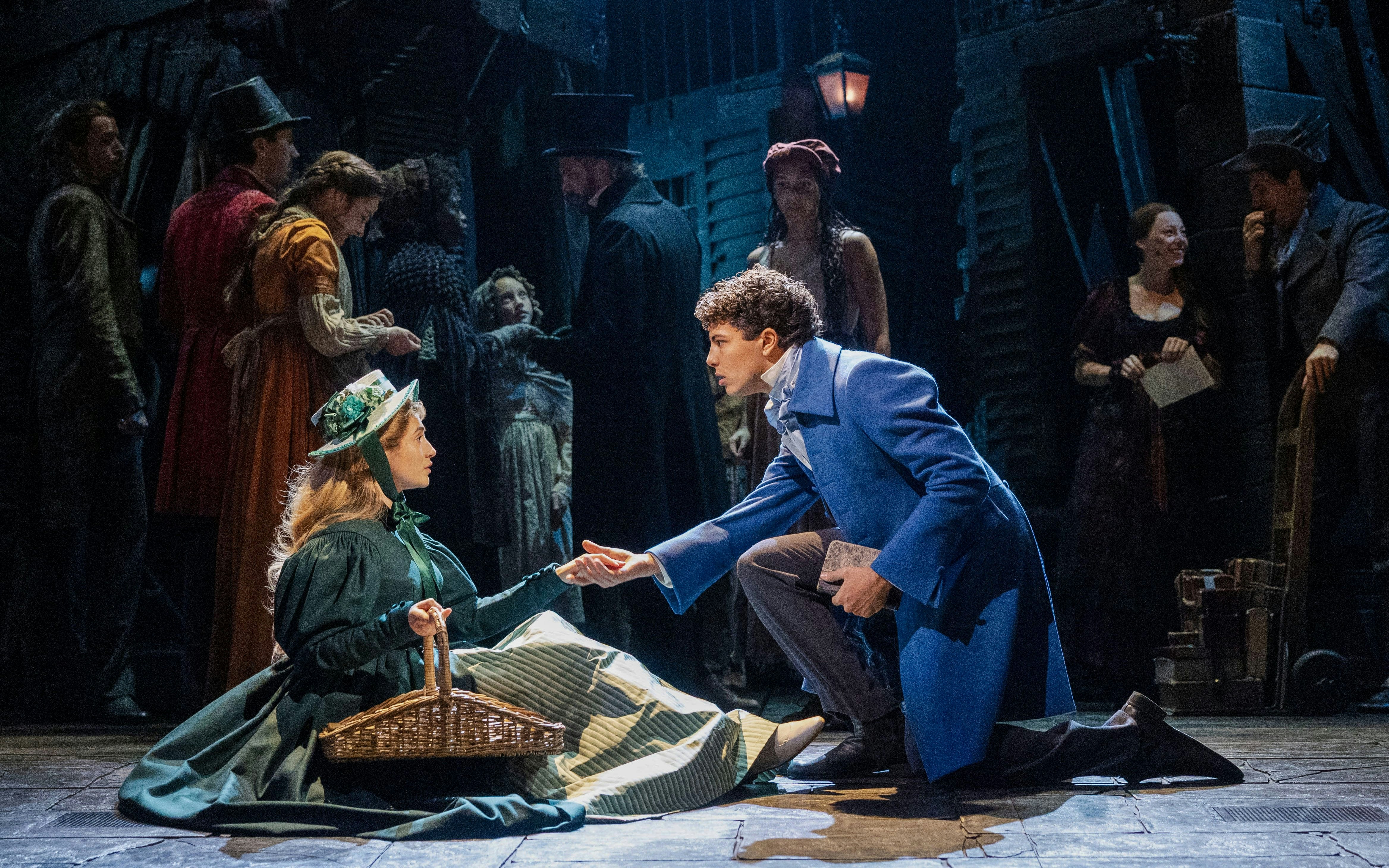 Les Misérables stage scene with actors in period costumes, one kneeling and offering a hand.