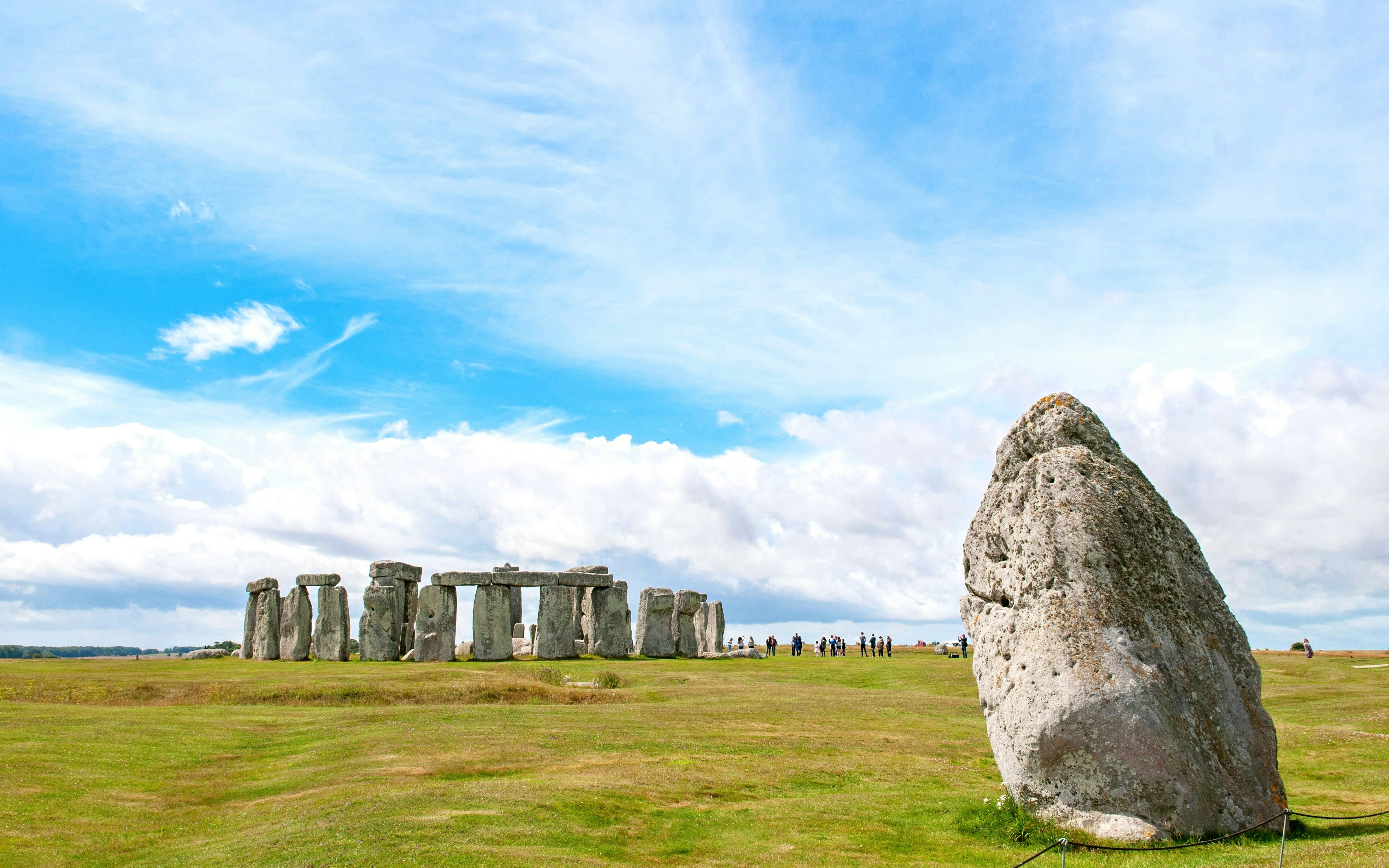 Heel Stone at Stonehenge with standing stones in the background under a blue sky.