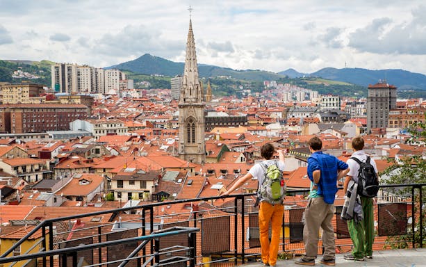 Tourists overlooking Bilbao cityscape with view of church spire and surrounding hills.