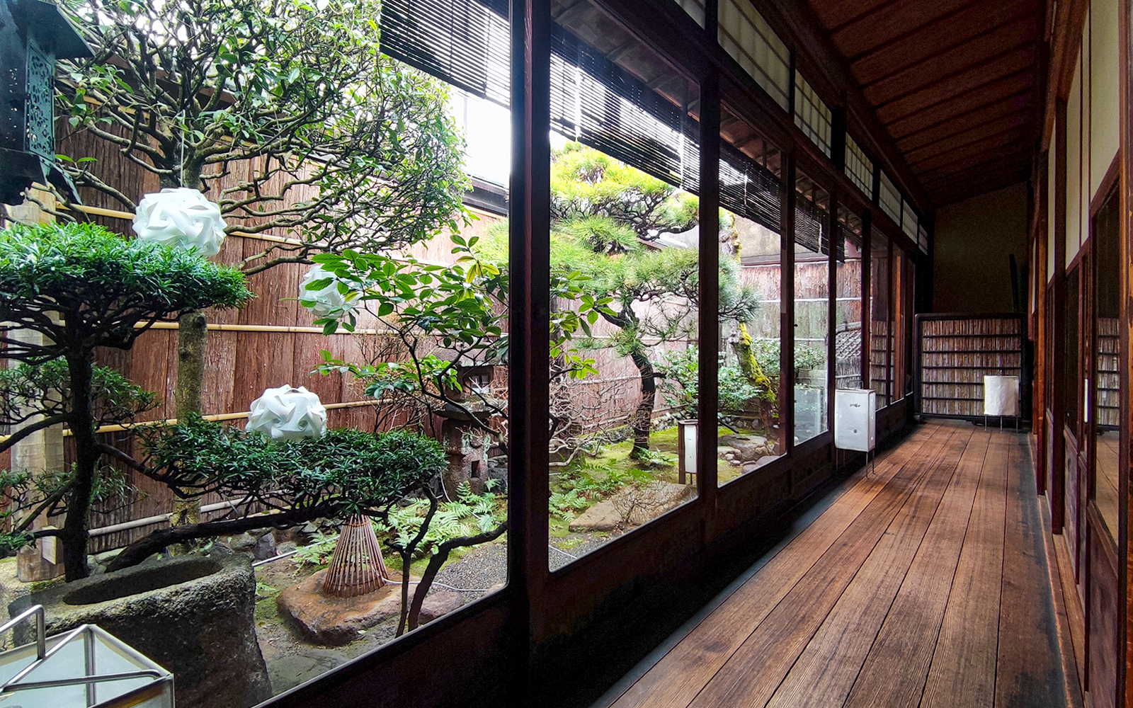 Kyoto machiya interior with traditional garden view, featuring bonsai trees and wooden walkway.