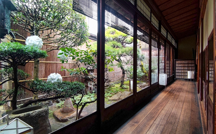 Kyoto machiya interior with traditional garden view, featuring bonsai trees and wooden walkway.