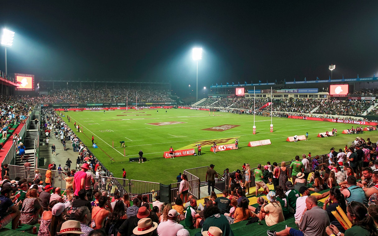 Crowd watching a rugby match at Emirates Dubai 7s stadium at night.