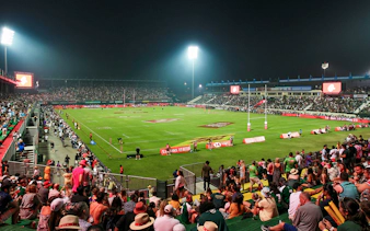 Crowd watching a rugby match at Emirates Dubai 7s stadium at night.