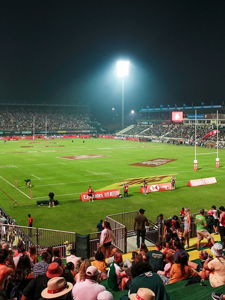Crowd watching a rugby match at Emirates Dubai 7s stadium at night.