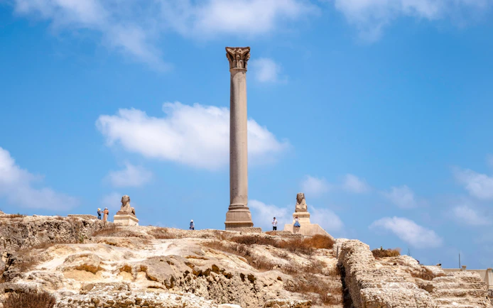 Pompey's Pillar in Alexandria, Egypt, with surrounding ruins and sphinx statues.