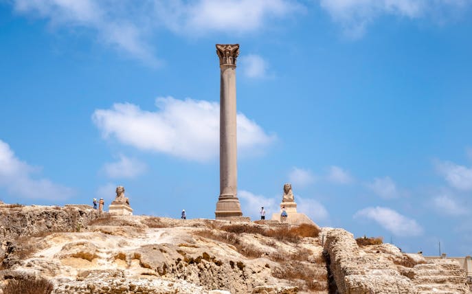 Pompey's Pillar in Alexandria, Egypt, with surrounding ruins and sphinx statues.