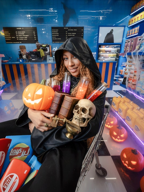 Woman in hooded cloak holding Halloween items at Enchanted Carnival's spooky shopping center.