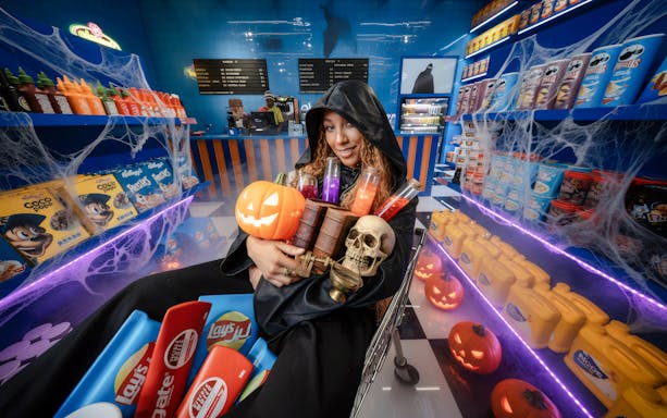 Woman in hooded cloak holding Halloween items at Enchanted Carnival's spooky shopping center.
