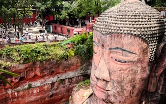 Leshan Giant Buddha with tourists in Chengdu, surrounded by greenery and red cliffs.
