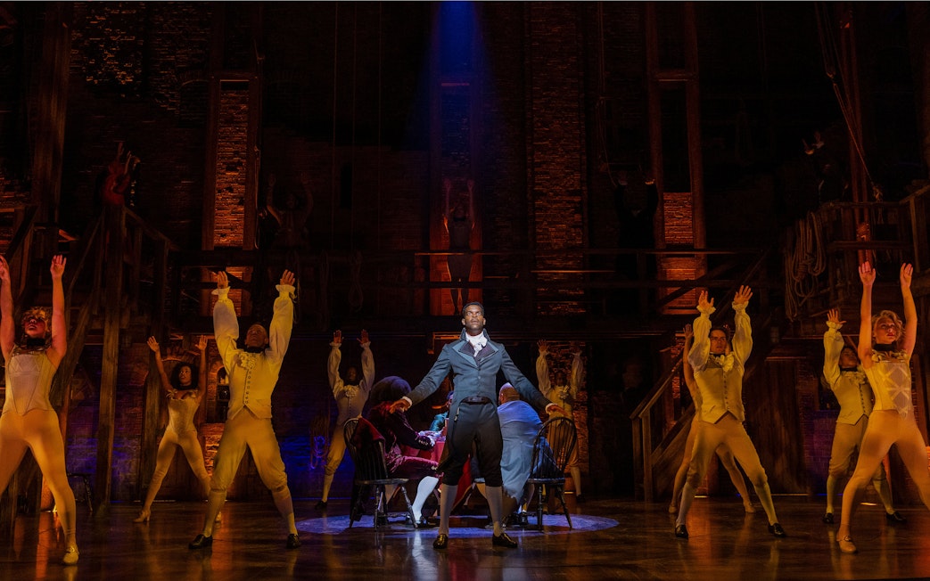 Cast performing a scene from Hamilton on stage with dramatic lighting.