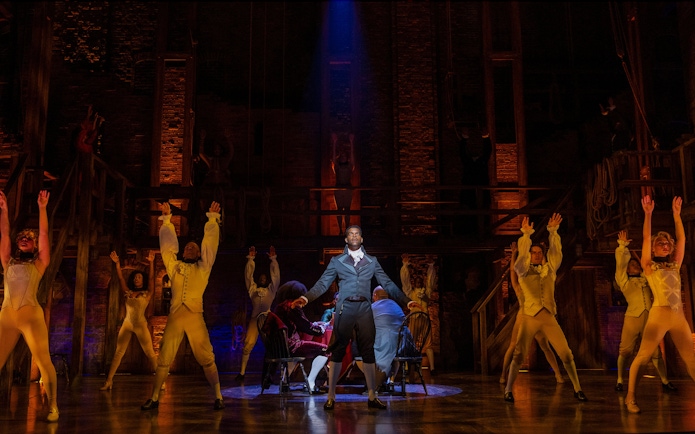 Cast performing a scene from Hamilton on stage with dramatic lighting.