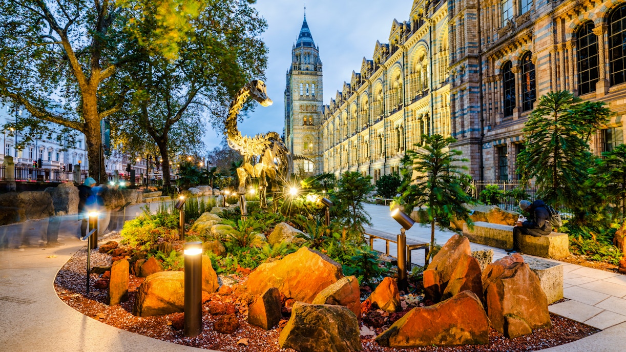 Dinosaur sculpture outside Natural History Museum, London, England, with illuminated facade.