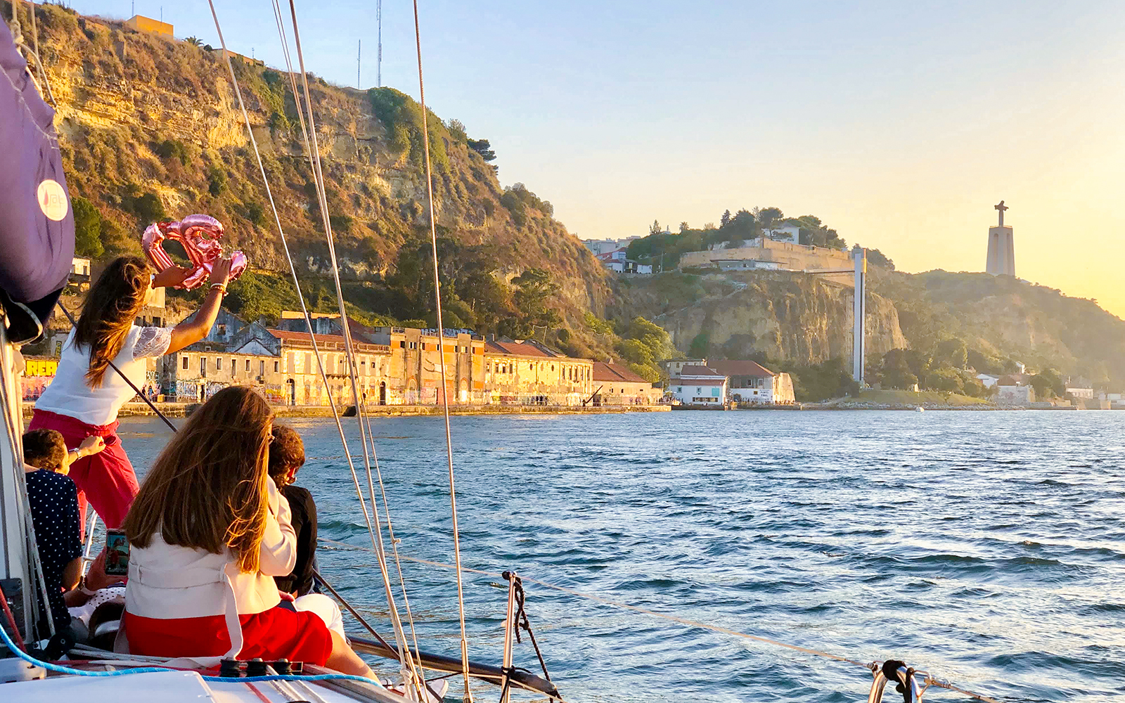 Tourists on a private boat enjoying Lisbon's waterfront view with Cristo Rei statue in the background.
