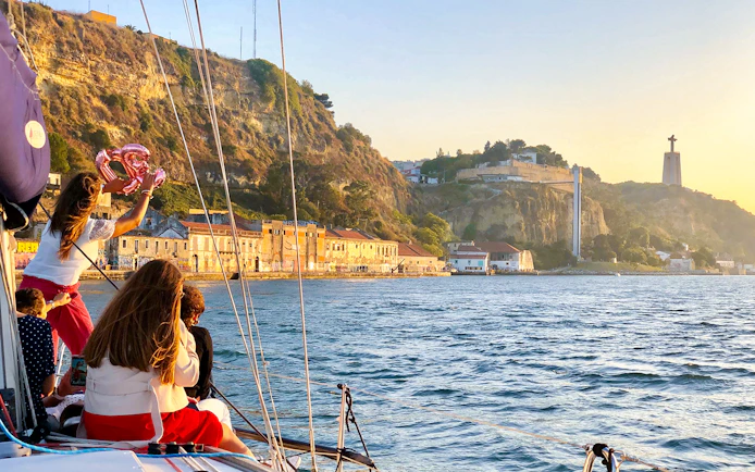 Tourists on a private boat enjoying Lisbon's waterfront view with Cristo Rei statue in the background.