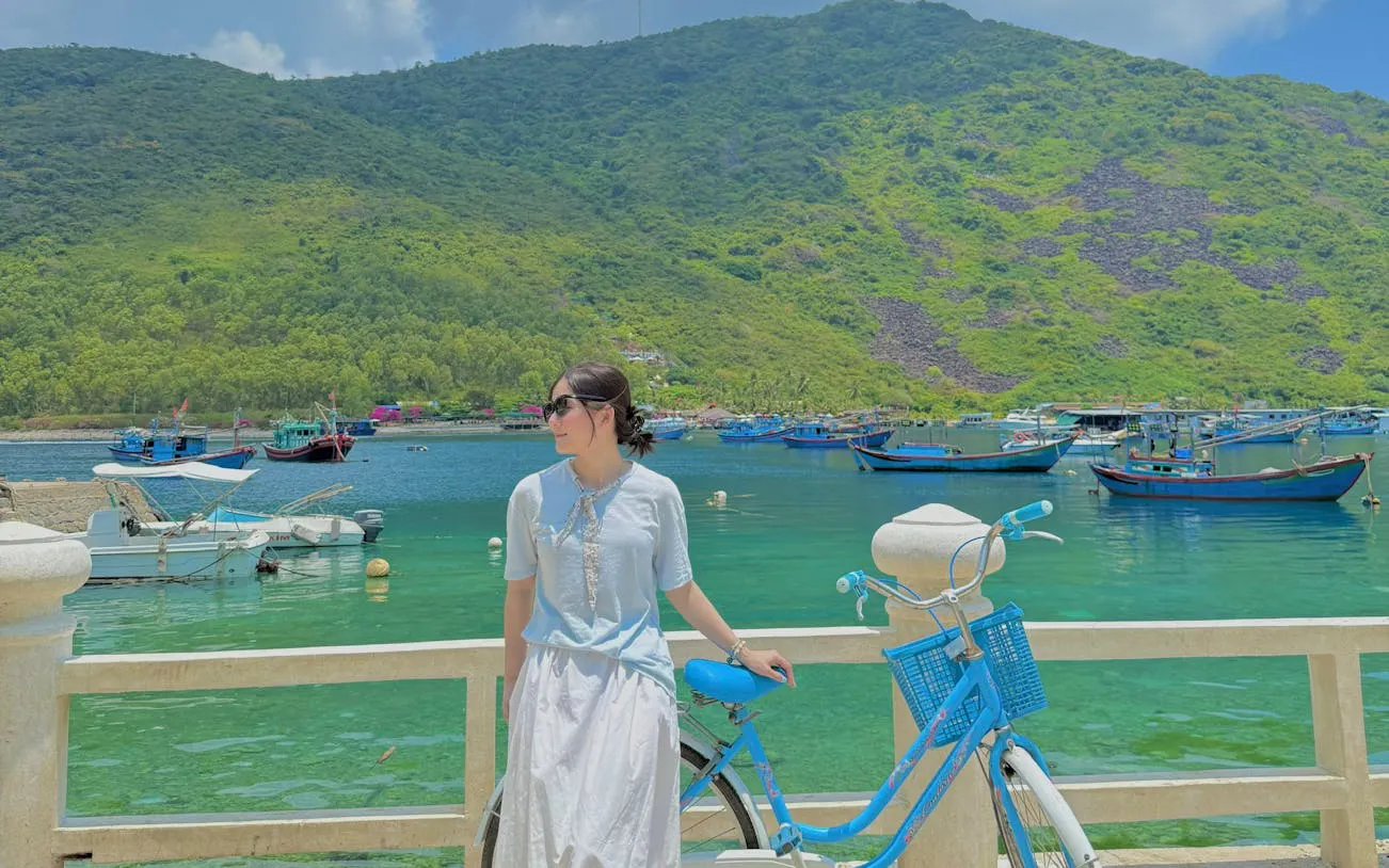 Woman with bicycle by Nha Trang's Robinson Beach, boats and green hills in background.