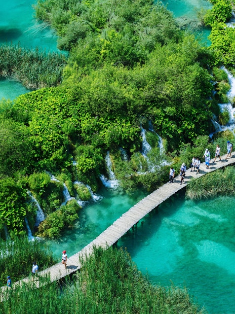 Visitors walking on a wooden path over turquoise lakes in Plitvice Lakes National Park, Croatia.
