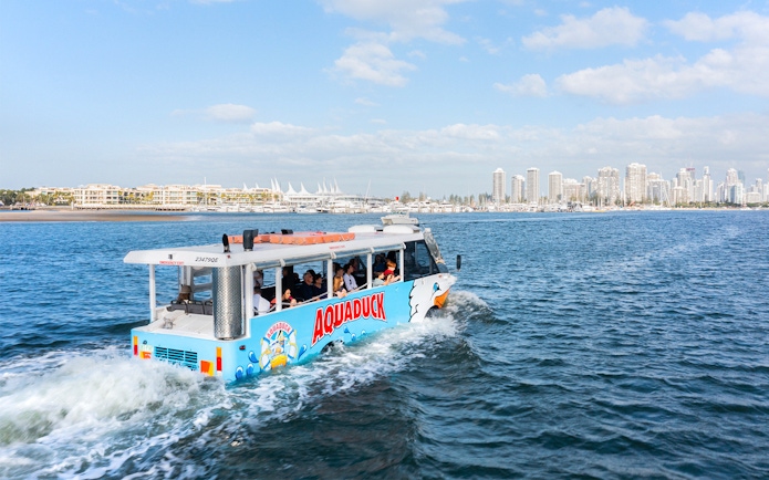 Aquaduck tour vehicle cruising on a river with city skyline in the background.