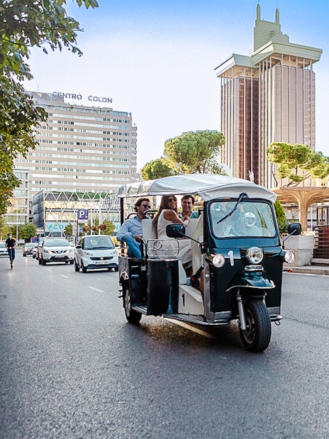 Eco Tuk Tuk driving through Madrid city street with modern buildings in the background.