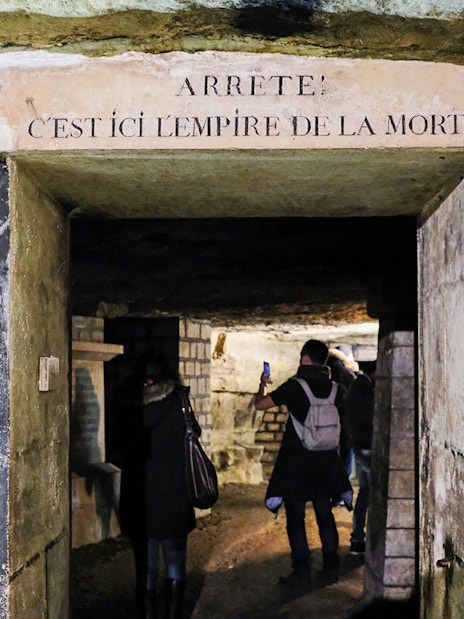 Visitors exploring the entrance of the Paris Catacombs with a sign above.