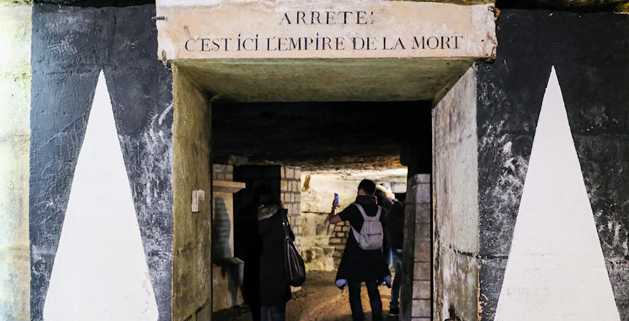 Visitors exploring the entrance of the Paris Catacombs with a sign above.