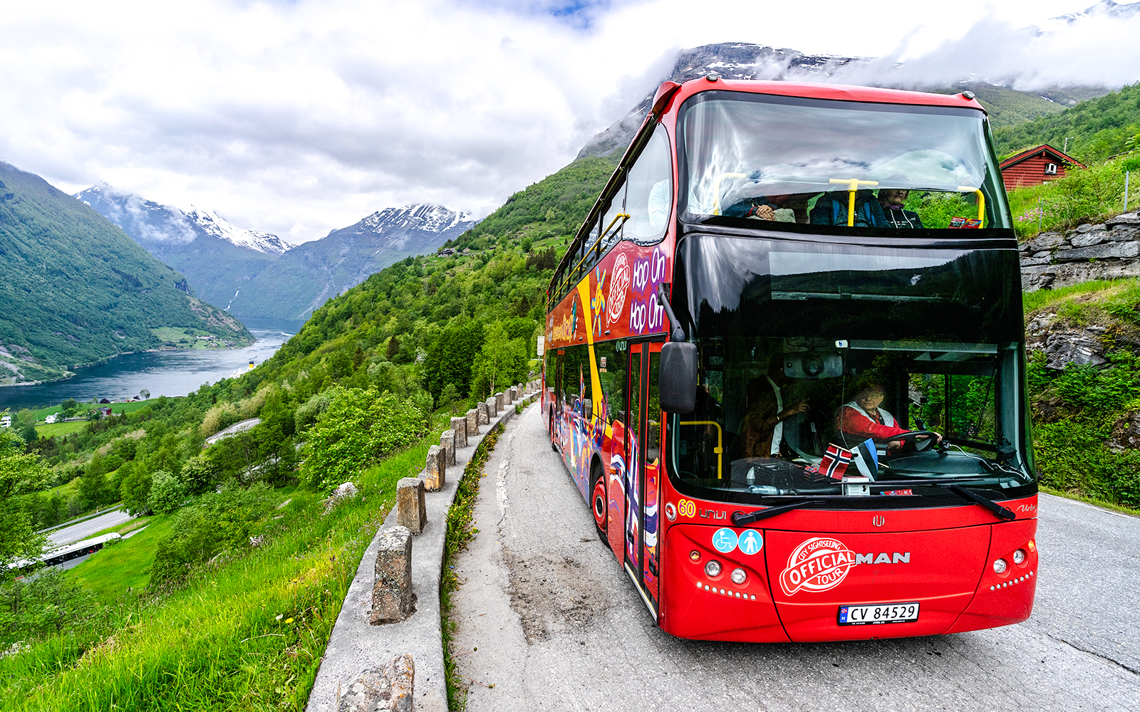 Red double-decker bus on Geiranger Hop-on Hop-off tour with fjord and mountains in background.