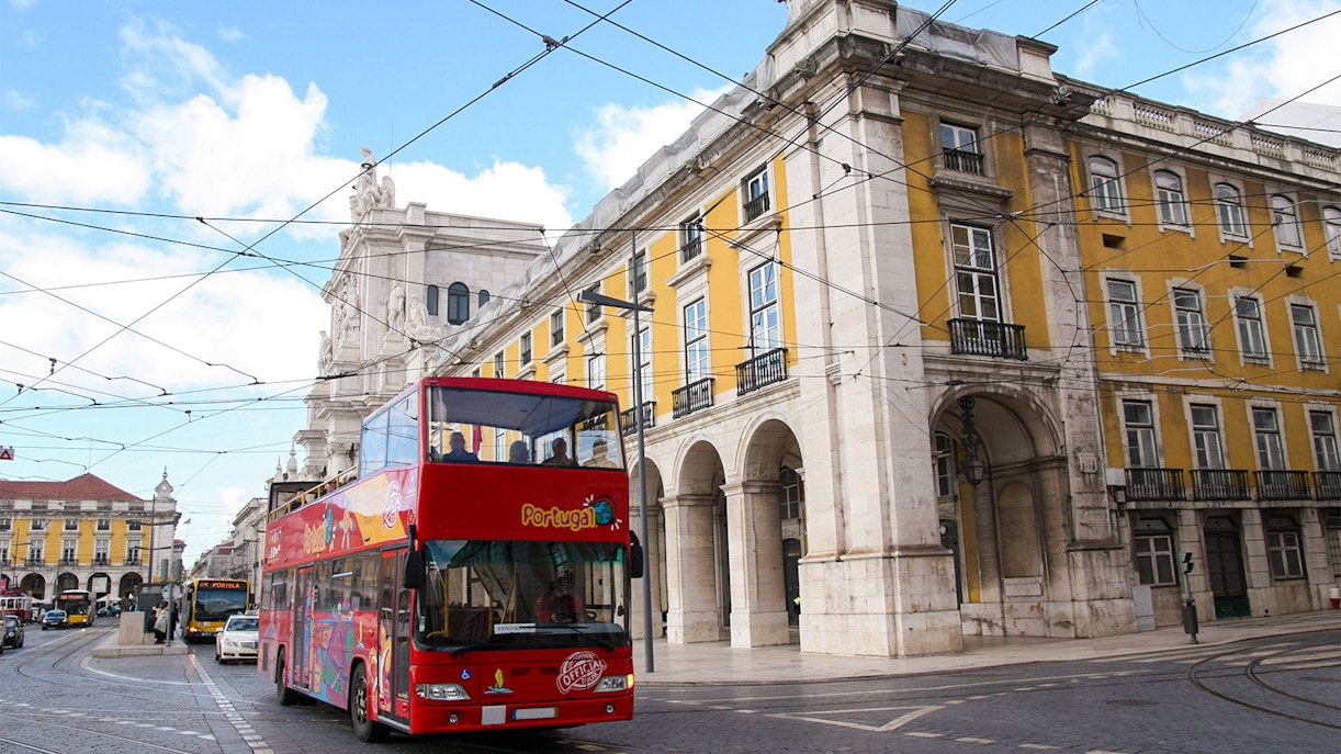 Red double-decker tour bus in Lisbon near historic yellow building, en route to Monserrate Palace & Park.