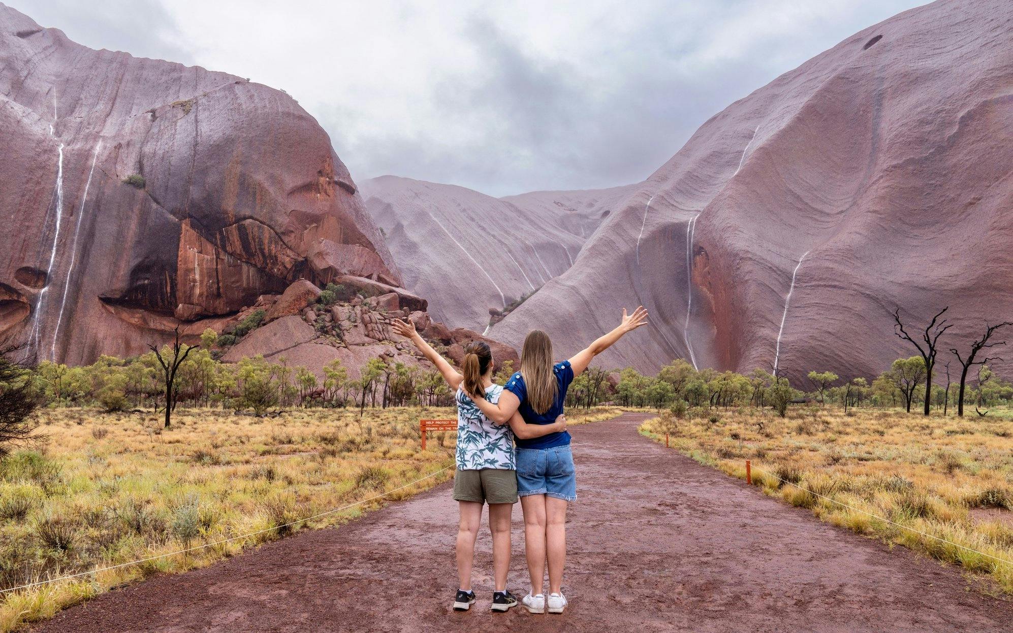 Two people embracing and facing Uluru rock formations on an Outback camping adventure.