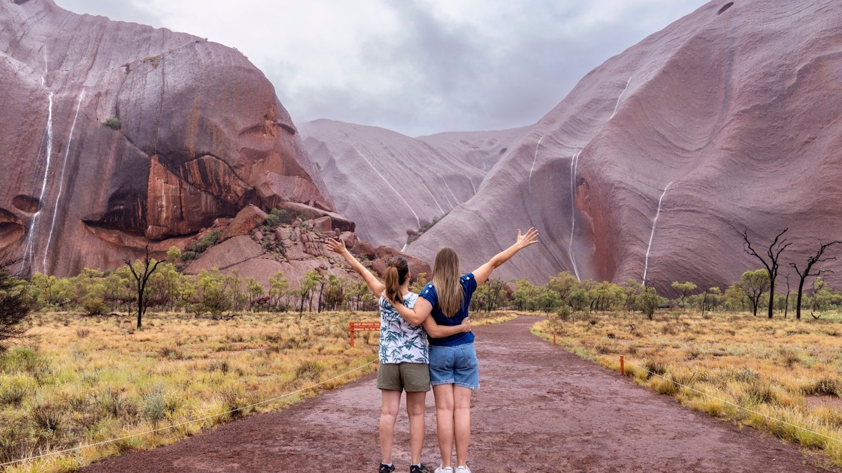 Two people embracing and facing Uluru rock formations on an Outback camping adventure.