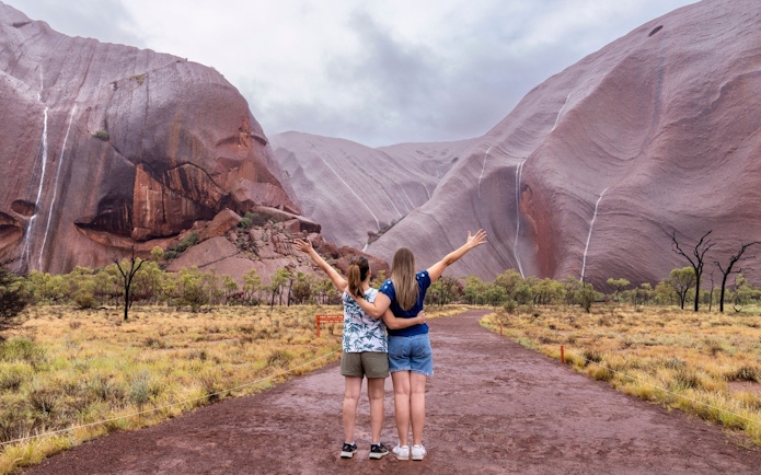 Two people embracing and facing Uluru rock formations on an Outback camping adventure.