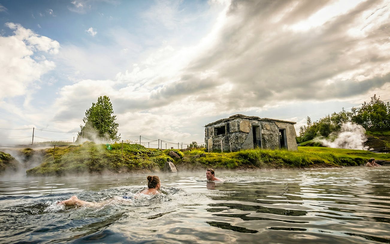 Guests swimming in Secret Lagoon with steam rising and a stone building nearby.