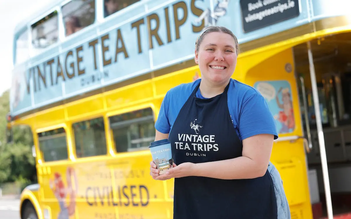 Smiling person holding a cup in front of a yellow Vintage Tea Trips bus in Dublin.