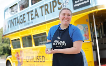 Smiling person holding a cup in front of a yellow Vintage Tea Trips bus in Dublin.