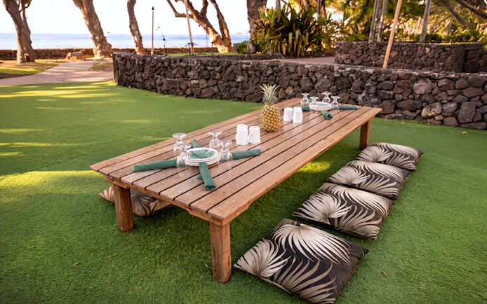 Outdoor seating setup at Old Lahaina Luau with wooden table, cushions, and ocean view.