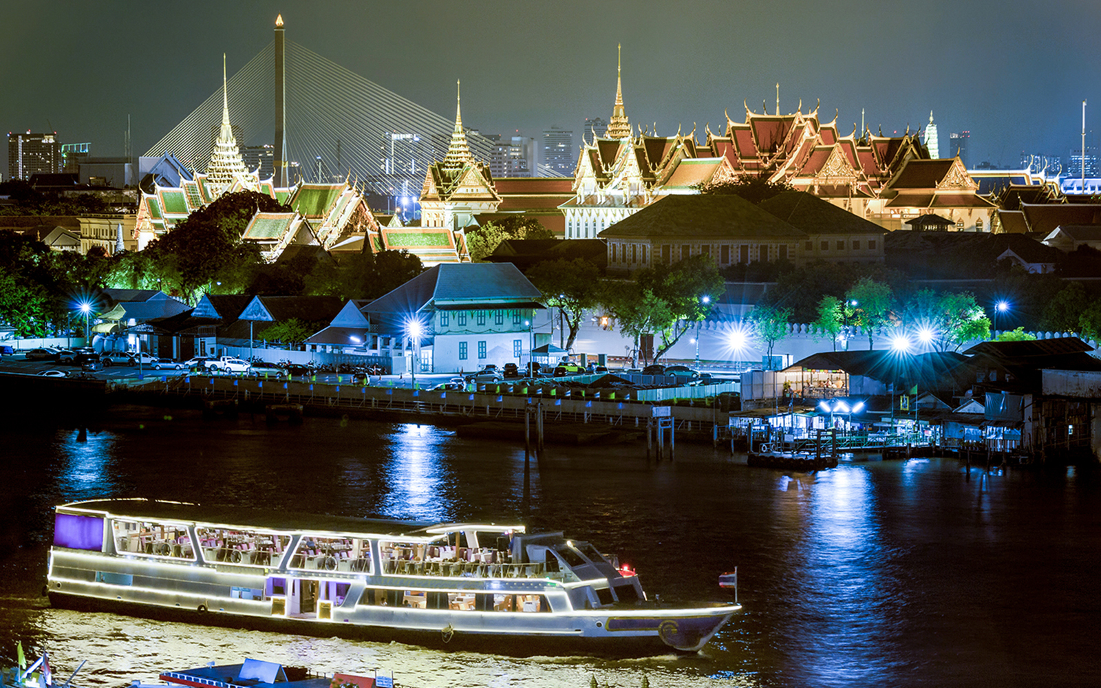 Cruise on Chao Phraya River with illuminated Grand Palace and Rama VIII Bridge in Bangkok.