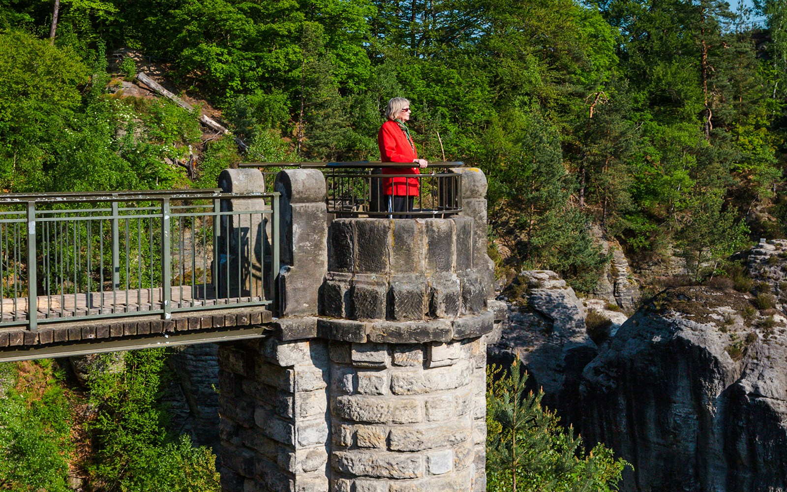 Woman standing at viewpoint on Bastion Bridge, surrounded by lush greenery and rocky cliffs.
