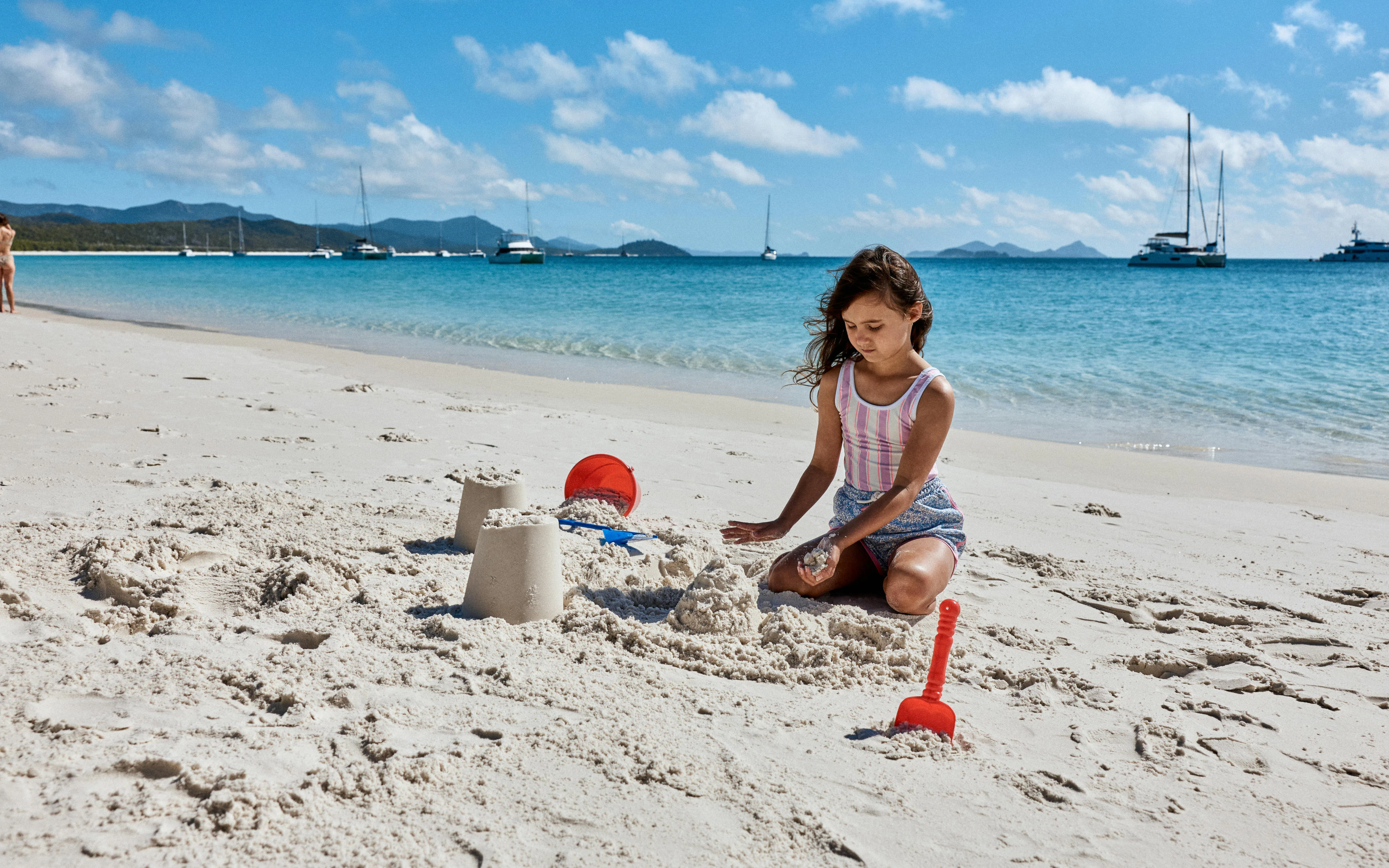 Child building sandcastles on Whitehaven Beach, Whitsundays, with yachts in the background.