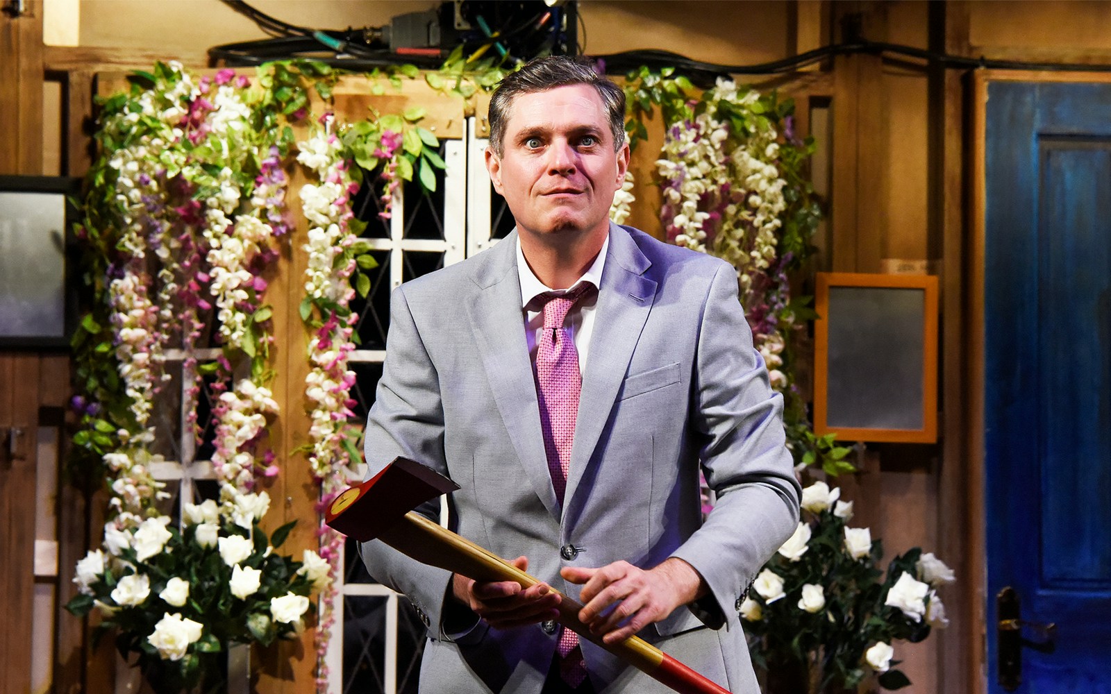 Man in a suit holding an axe on stage with floral backdrop, "Noises Off" play.